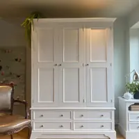 Cream painted Carlton triple wardrobe with three panelled doors above four drawers on a shaped plinth, with a trailing fern on top and a leather armchair alongside