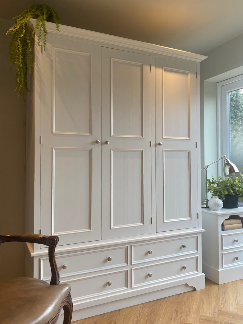 Angled full view of the pale grey painted three door wardrobe with four drawers beneath, round nickel knobs and a trailing fern on top, beside a window with a lamp and plant