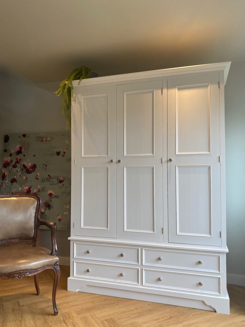 Three-quarter view of the wardrobe showing its moulded cornice, three panelled doors and four base drawers, with a floral canvas and a carved leather chair to the left