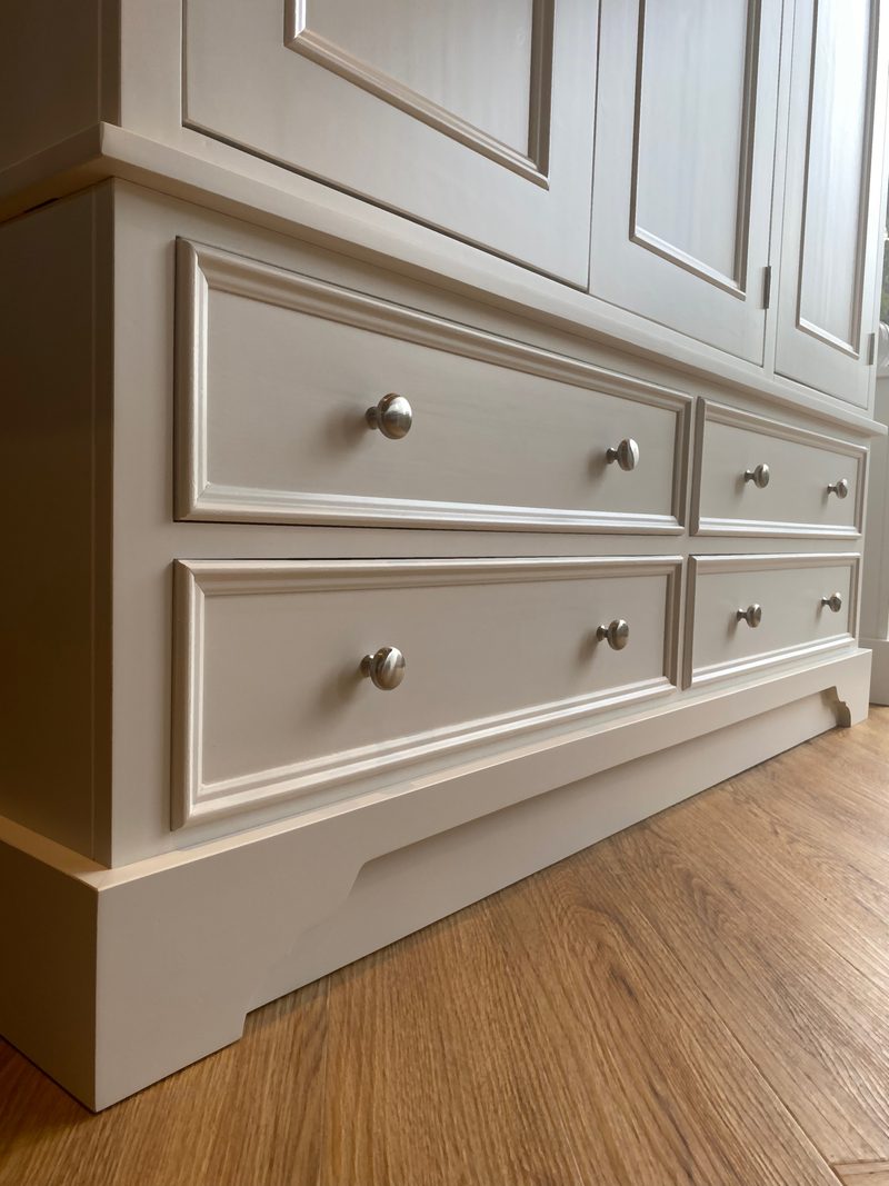 Angled view of the four base drawers beneath the panelled doors, showing the moulded fronts, paired round nickel knobs and the shaped plinth standing on oak flooring