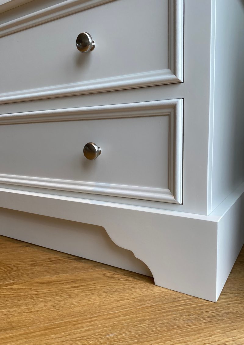 Close corner detail of two stacked drawers and the shaped bracket plinth, showing the moulded drawer frames, the round nickel knob on each front and the light oak floor below