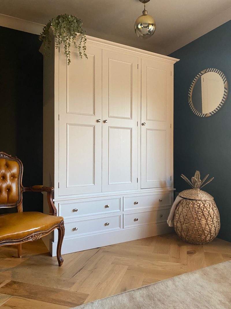 Cream painted Chellington triple wardrobe with three panelled doors above four drawers, against a dark teal wall on herringbone oak flooring with a tan buttoned chair alongside