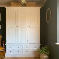 Straight-on view of the cream painted wardrobe with three panelled doors above four base drawers, brushed nickel knobs, wicker baskets on top and a tan leather chair against a dark teal wall