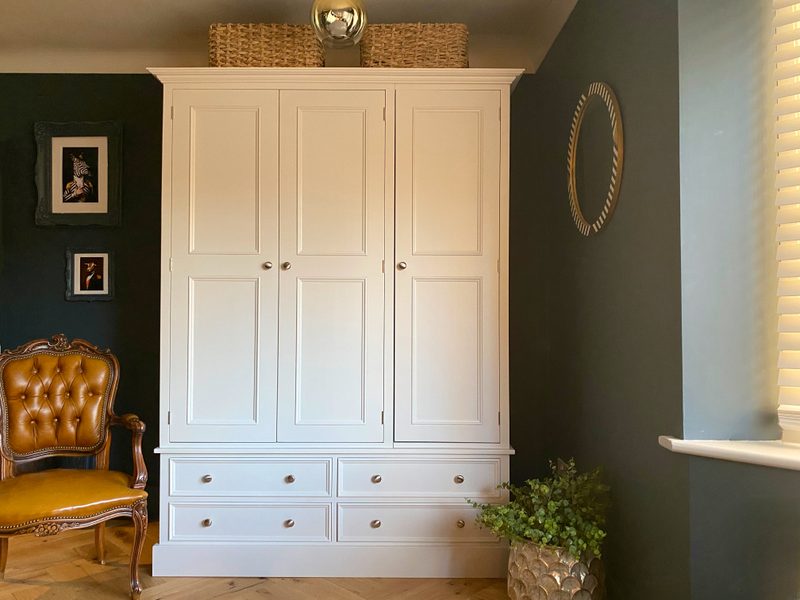 Straight-on view of the cream painted wardrobe with three panelled doors above four base drawers, brushed nickel knobs, wicker baskets on top and a tan leather chair against a dark teal wall