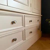 Low angled close view along the base of the wardrobe showing four cream painted drawers with brushed nickel knobs, the moulded plinth and the herringbone oak floor beside a dark wall