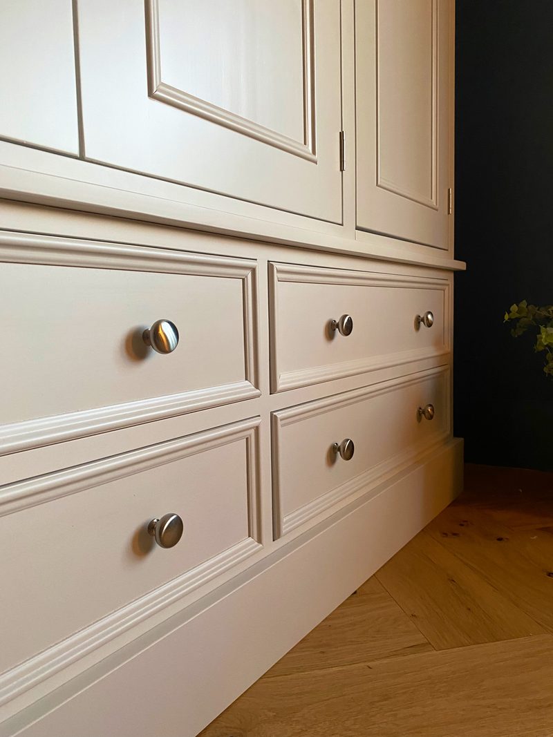 Low angled close view along the base of the wardrobe showing four cream painted drawers with brushed nickel knobs, the moulded plinth and the herringbone oak floor beside a dark wall