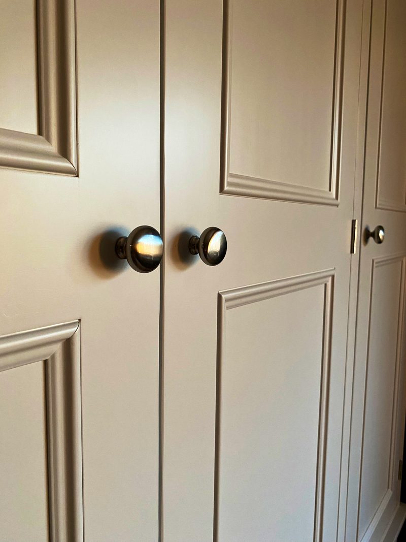 Close view of the meeting point of two panelled wardrobe doors, showing a pair of round brushed nickel knobs, the moulded panel edges and a third door knob further along the front