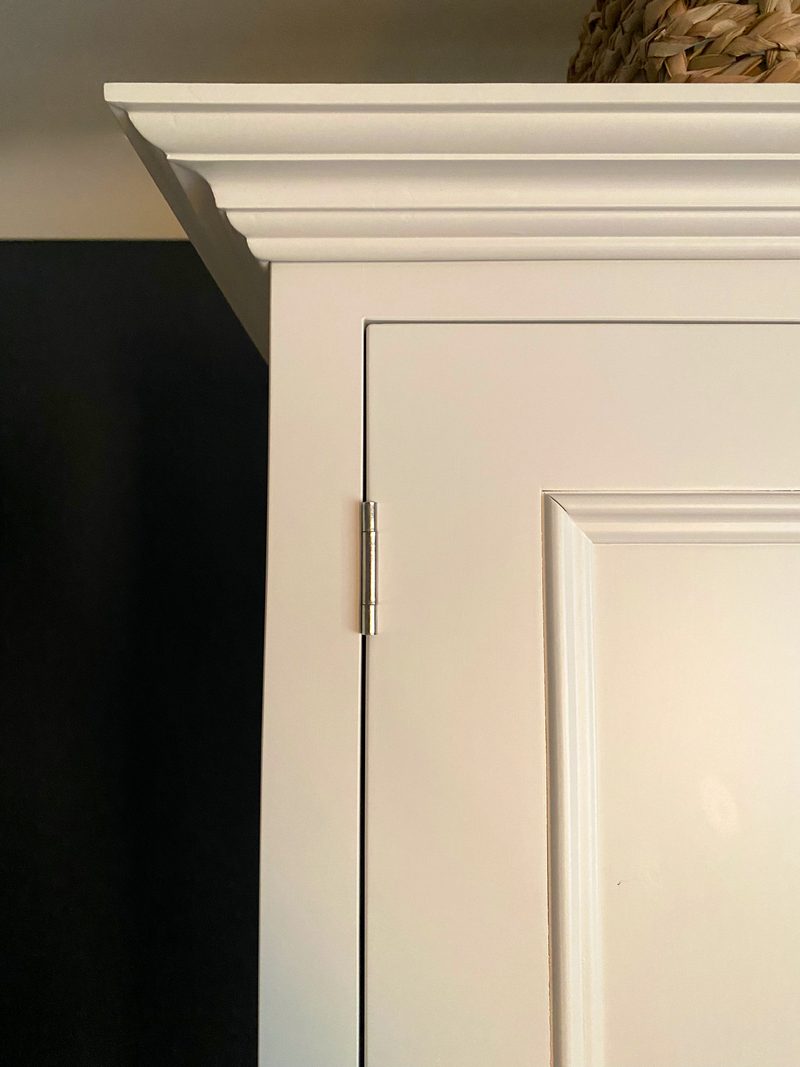Detail of the top corner of the wardrobe, showing the stepped cornice moulding, a slim metal barrel hinge, the top of a panelled door and a woven basket resting above it