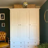 Full front view of the wardrobe with three panelled doors above four drawers, two wicker baskets and a brass globe pendant overhead, framed prints and a tan leather chair alongside