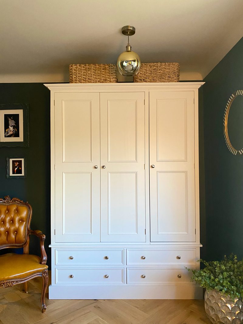Full front view of the wardrobe with three panelled doors above four drawers, two wicker baskets and a brass globe pendant overhead, framed prints and a tan leather chair alongside