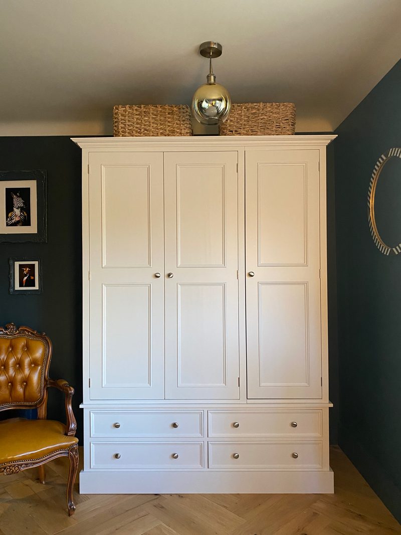 Wider front view of the wardrobe standing on a herringbone oak floor, its four base drawers and brushed nickel knobs clear, with a round striped mirror on the dark green wall to the right
