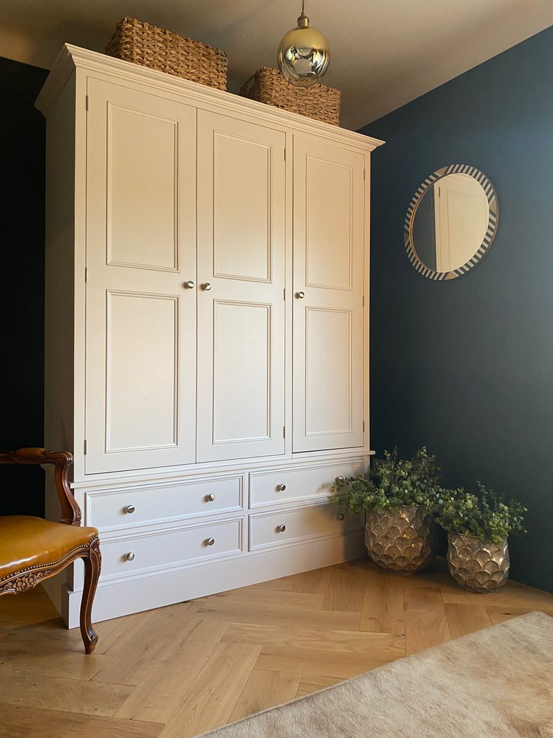 Three-quarter view from the left showing the depth of the wardrobe carcass, the cornice topped with woven baskets and two metallic planters of trailing greenery on the herringbone floor