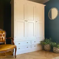 Angled room view of the wardrobe centred between a buttoned tan leather chair and two metallic planters, its cream doors and drawers set against a deep teal painted wall