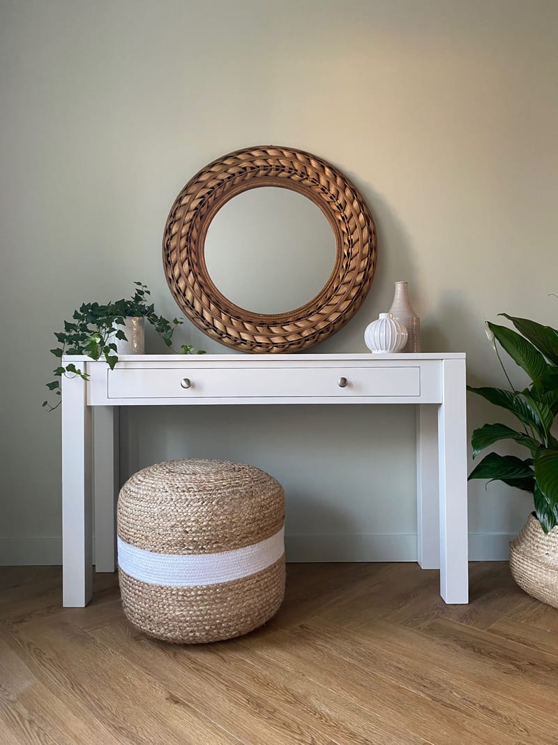 Straight-on view of the white painted console table with one long drawer and two brushed nickel knobs, styled beneath a round woven rattan mirror with a jute pouffe tucked below