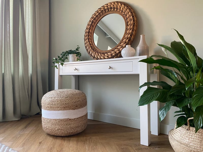 Angled landscape view of the console table beside sage green curtains, showing the straight square legs, the long single drawer with two nickel knobs and a jute pouffe tucked underneath