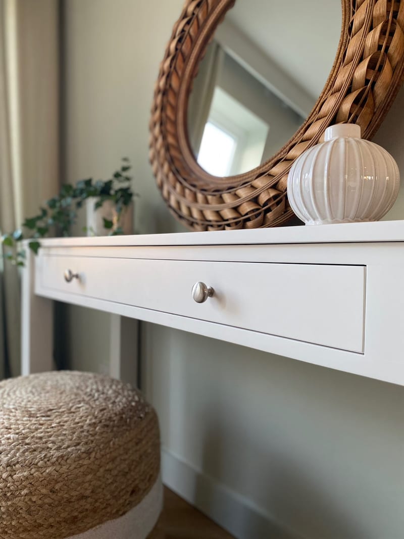 Close view along the top edge and drawer front of the console table, showing the crisp white paint, two round brushed nickel knobs and a ribbed white vase in front of the rattan mirror