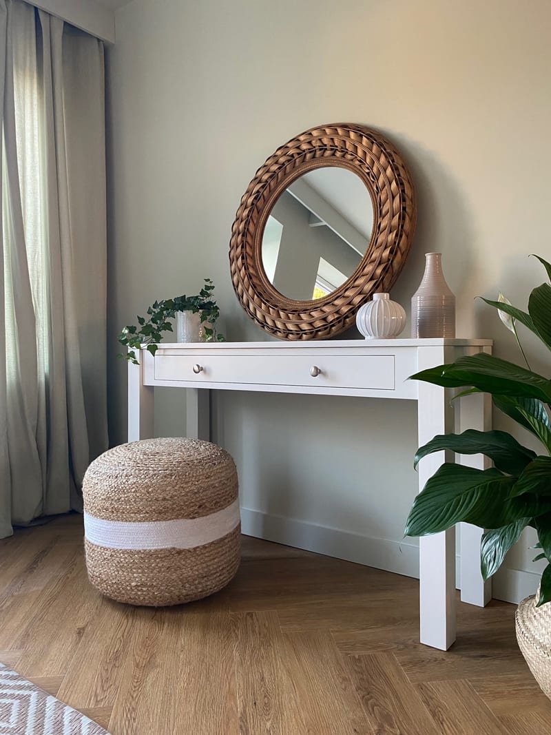 Portrait view of the console table against a soft green wall, with linen curtains to one side, a peace lily to the other and a striped jute pouffe on the herringbone wood floor