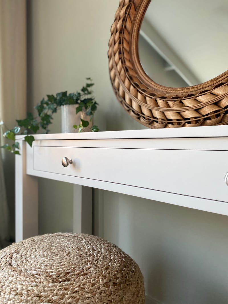Close view of the left end of the console table showing the corner of the white painted top, a square leg, a round brushed nickel knob and a pot of ivy beside the woven rattan mirror