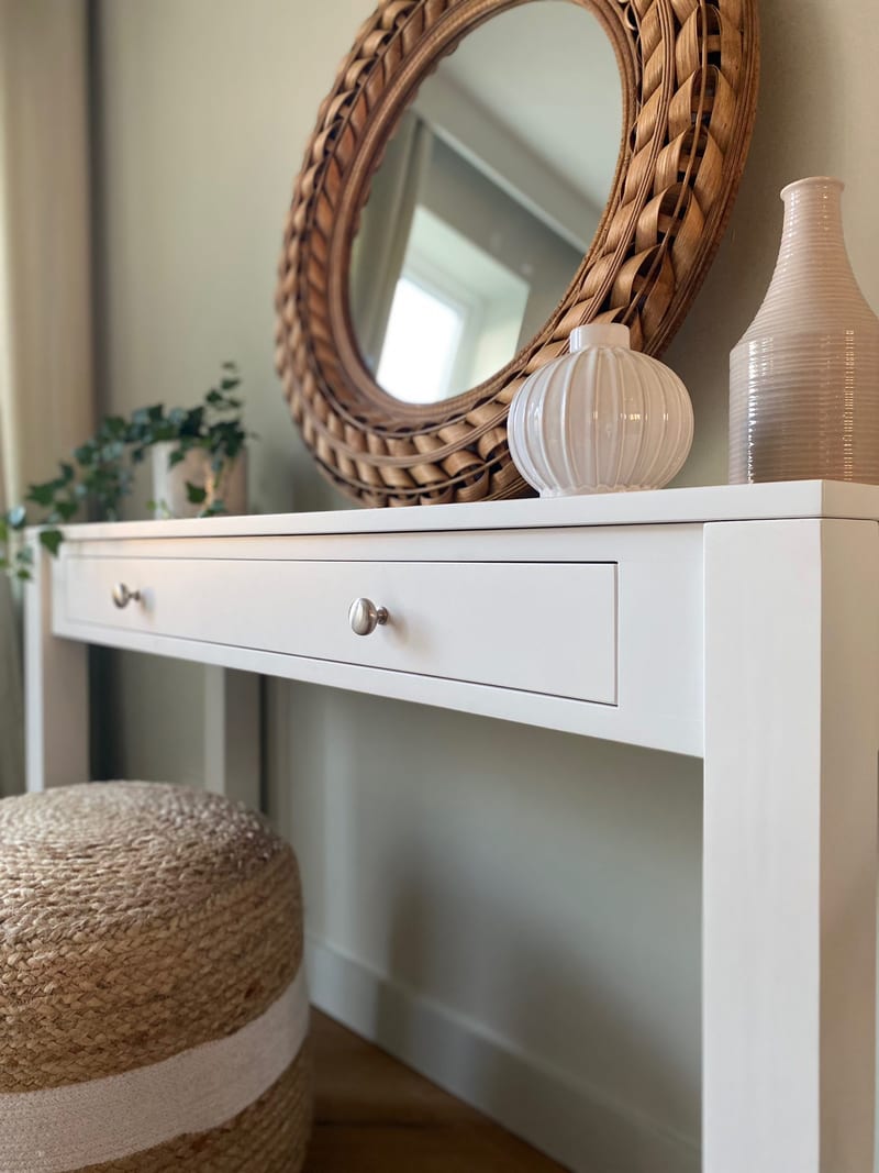 Close view of the right end of the table top with the drawer front and two knobs, a tall tapered vase and a small ribbed vase below the round rattan mirror on the pale green wall