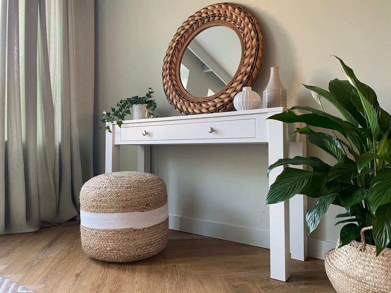 Wide landscape view of the console table with its top styled with ivy, a ribbed vase and a tall vase, the round rattan mirror above and a jute pouffe on the wood floor by a curtain
