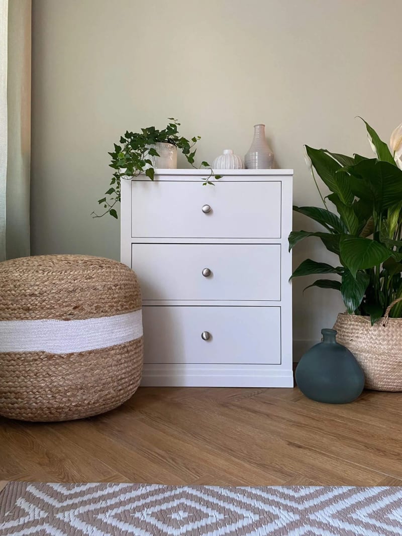 White painted Colmworth three drawer chest with round knobs, styled with trailing ivy and ribbed vases, beside a jute pouffe and a peace lily in a seagrass basket