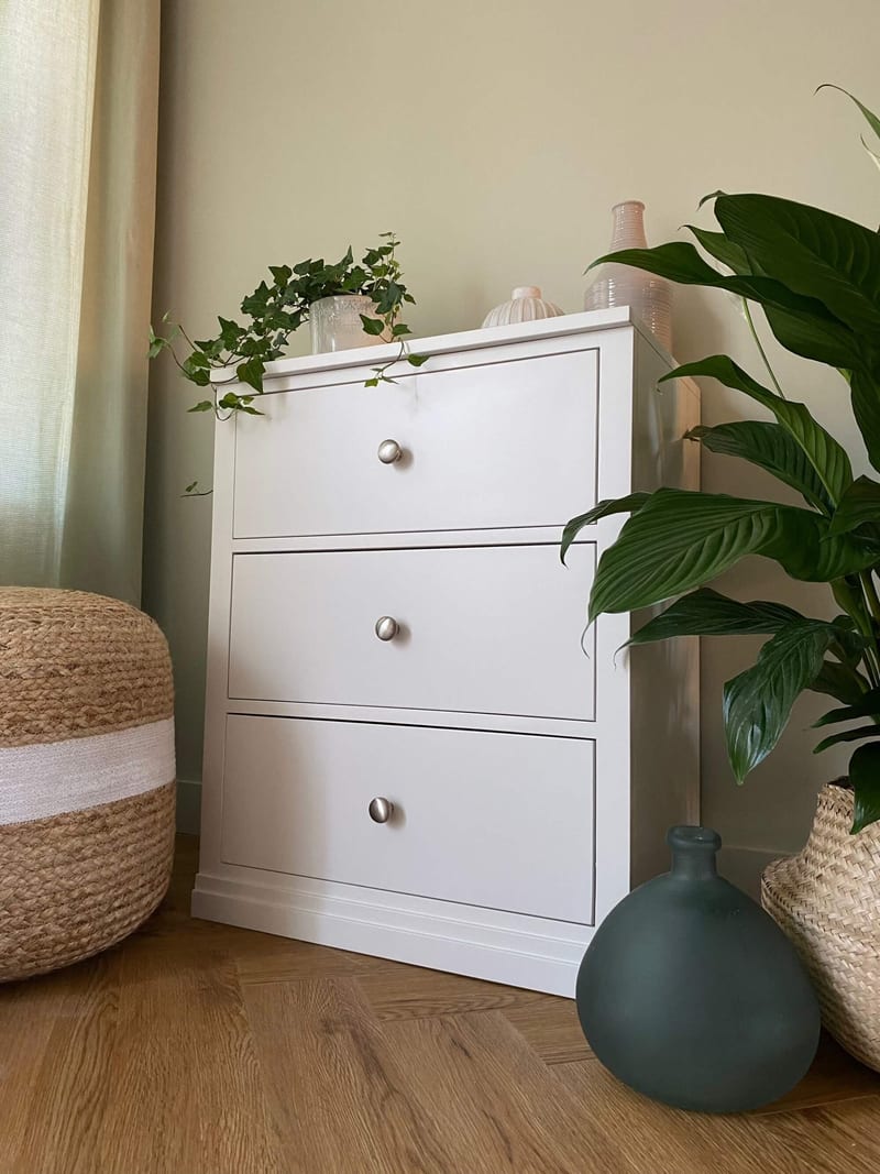 Angled view of the white painted three-drawer chest with a single brushed nickel knob on each drawer, trailing ivy on top and a teal glass vase beside it on the herringbone floor
