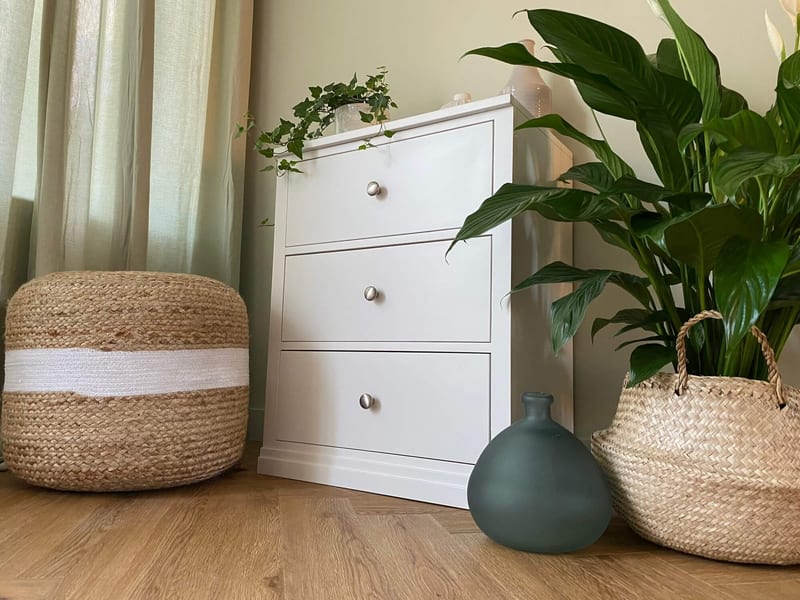 Landscape view of the three-drawer chest in a corner beside pale curtains, with a jute pouffe to the left and a peace lily in a seagrass basket and a teal vase to the right