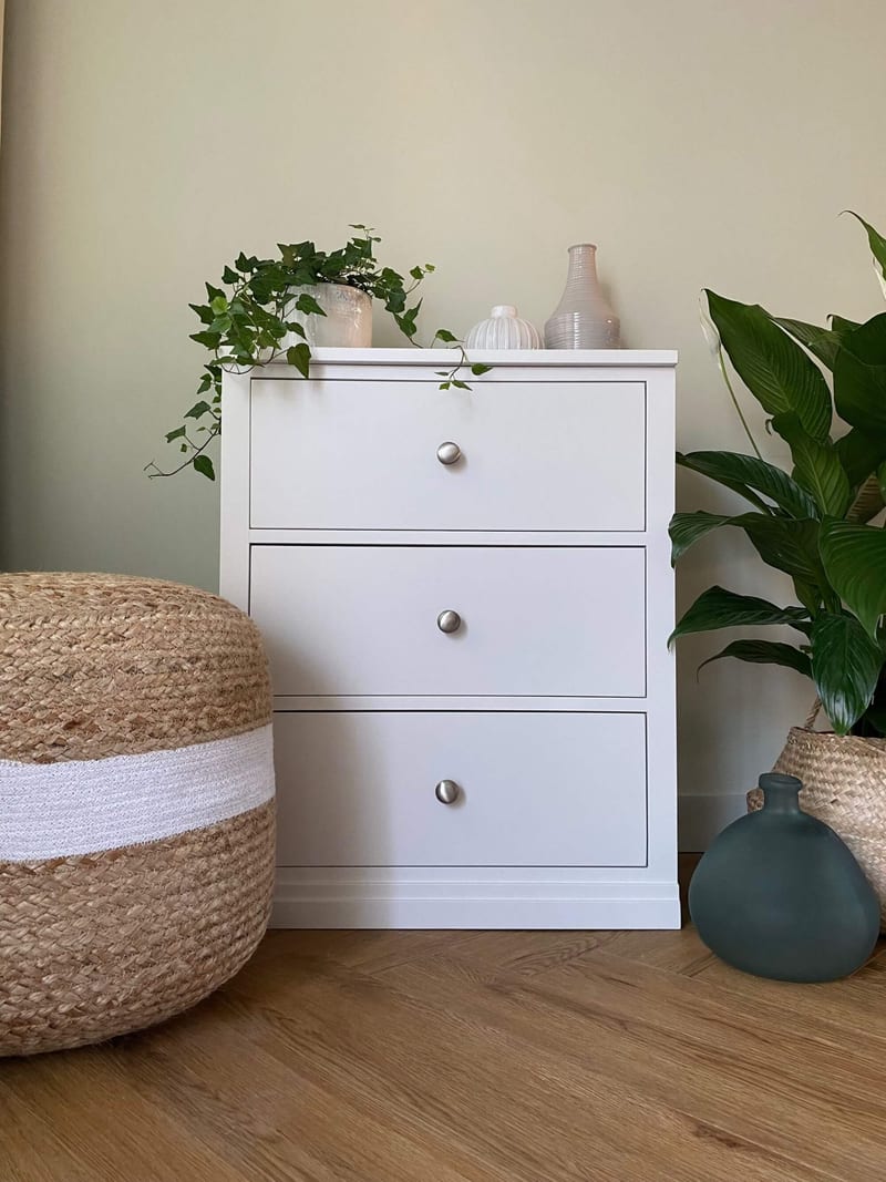 Front view of the chest showing three deep drawers, the moulded top edge and plinth, with ivy and two pale vases displayed above and a woven jute pouffe standing alongside