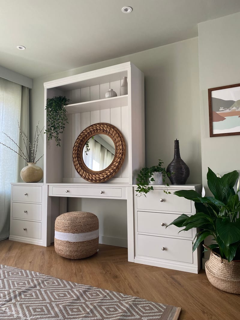 Angled view of the painted dressing table with its open display shelf, tongue-and-groove back panel and round woven rattan mirror, set between two flanking three-drawer chests