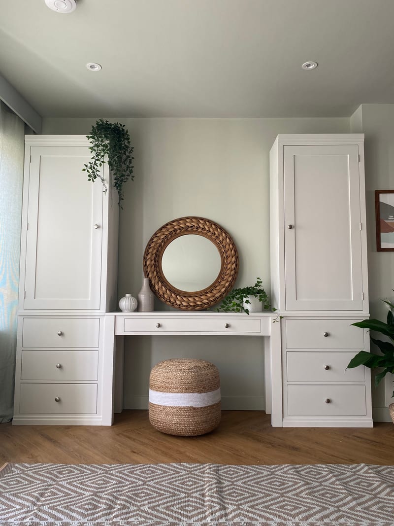 White painted Colmworth dressing table between two tall cupboards over three drawer chests, with a round rattan mirror above and a jute pouffe beneath
