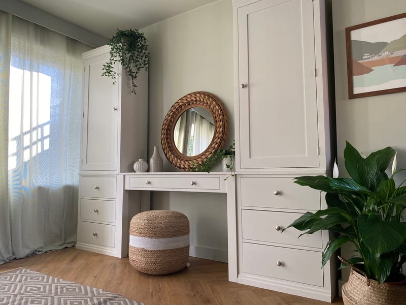 Wide landscape view of the dressing table lit by a sunlit curtained window on the left, with the jute pouffe tucked under the desk and a peace lily in a woven basket at the right