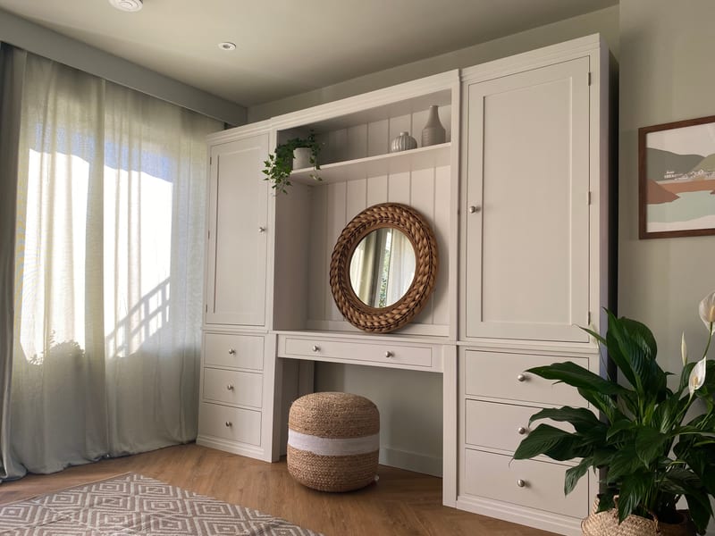 Landscape three-quarter view of the built-in dressing table between two tall cupboards, with sunlight through sage linen curtains on the left and a peace lily on the right