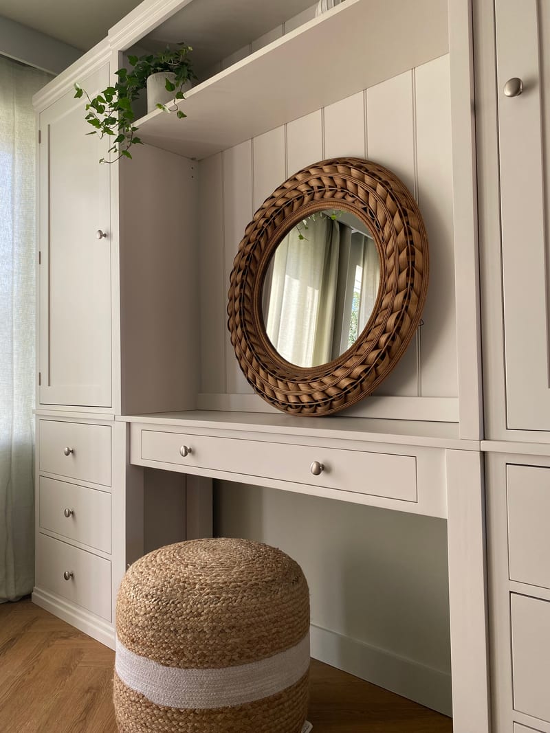 Close view of the round woven rattan mirror against the tongue-and-groove back panel, with a potted ivy on the shelf above and the desk drawer and brushed nickel knobs below
