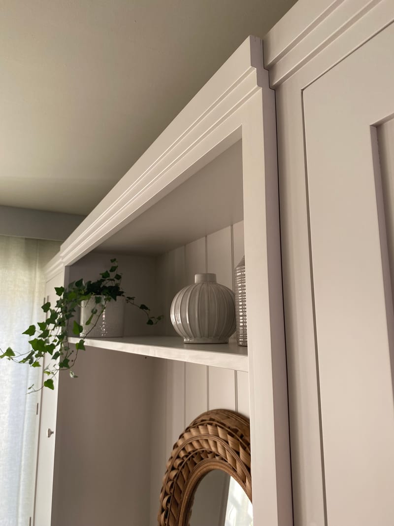 Close view of the moulded cornice and open display shelf, showing the tongue-and-groove back boards, a ribbed white vase, trailing ivy and the top of the rattan mirror below