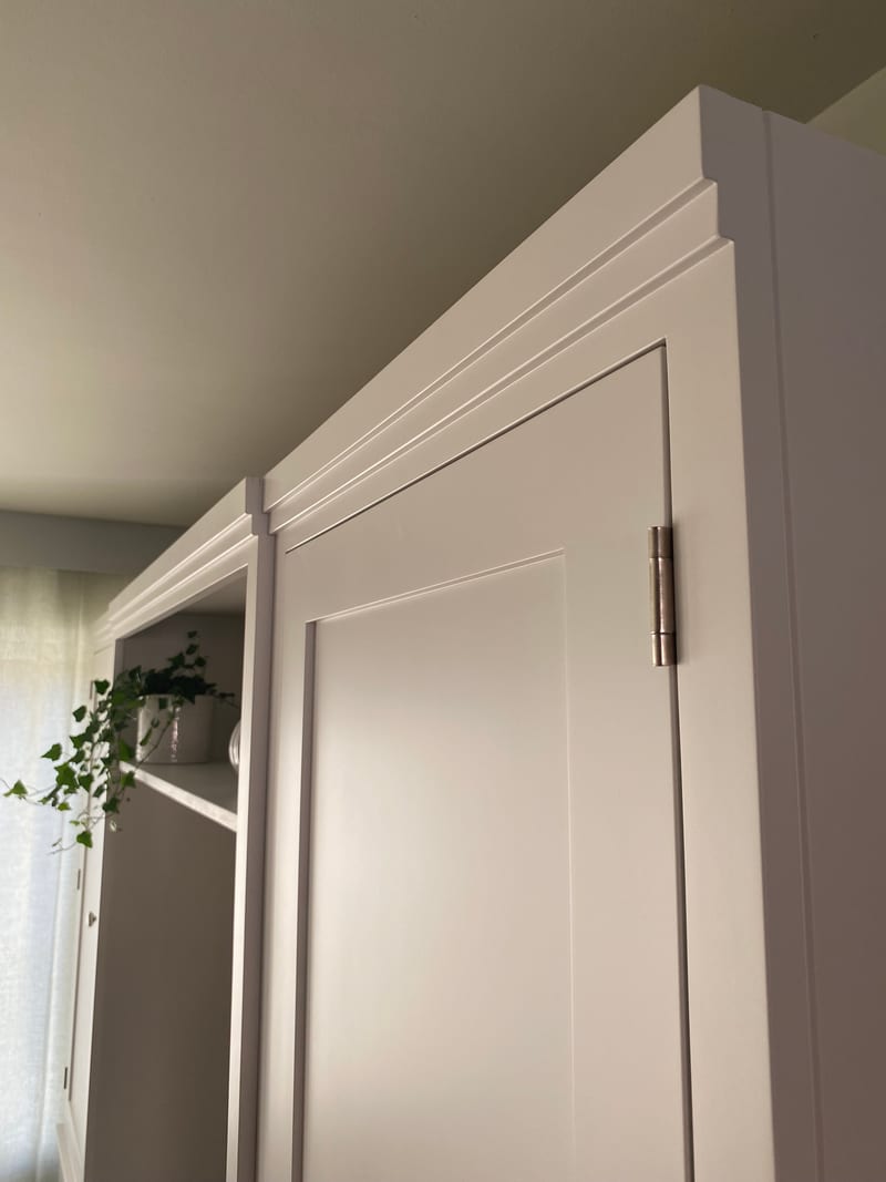 Detail of the top corner of the tall cupboard, showing the stepped cornice moulding, the recessed door panel and a brushed steel butt hinge against the pale painted ceiling
