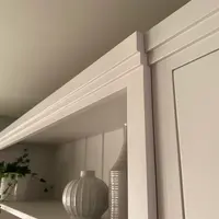Landscape detail of the moulded cornice above the display shelf, with a ribbed round vase and a tall ribbed bottle catching the light and the cupboard door frame to the right