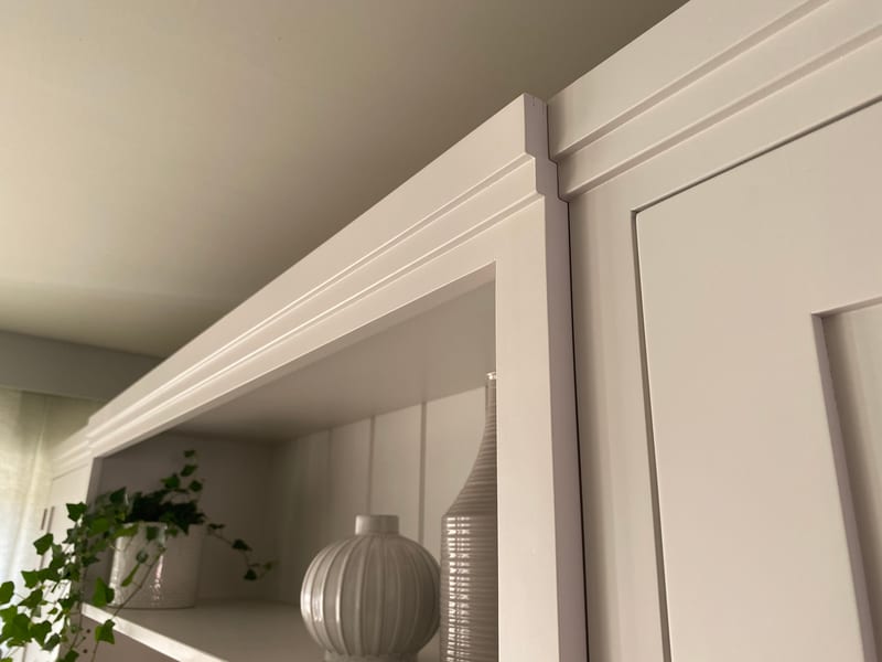 Landscape detail of the moulded cornice above the display shelf, with a ribbed round vase and a tall ribbed bottle catching the light and the cupboard door frame to the right