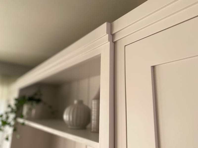 Very close shallow-focus detail of the corner post and cornice mouldings meeting the panelled cupboard door, with the shelf vases and trailing ivy blurred behind