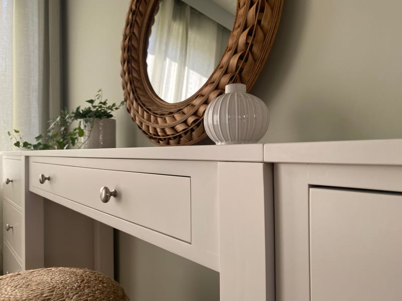 Close view along the top of the painted dressing table, showing the wide centre drawer with its brushed nickel knob, a ribbed white vase, trailing ivy and the woven rattan mirror behind