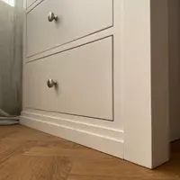 Low-angle view of the lower drawers of the dressing table pedestal, with brushed nickel knobs and stepped plinth, beside a sage linen curtain and herringbone oak flooring