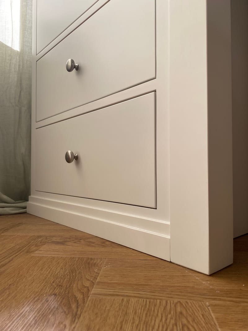 Low-angle view of the lower drawers of the dressing table pedestal, with brushed nickel knobs and stepped plinth, beside a sage linen curtain and herringbone oak flooring