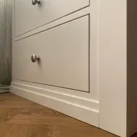 Corner detail of the painted pedestal at floor level, showing the smooth matt finish on the drawer fronts and the two-step moulded plinth meeting the herringbone oak floor