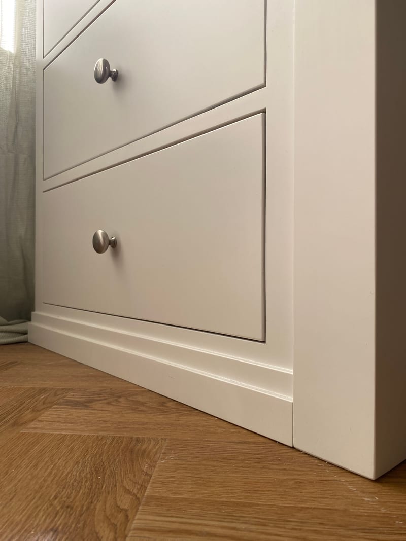 Corner detail of the painted pedestal at floor level, showing the smooth matt finish on the drawer fronts and the two-step moulded plinth meeting the herringbone oak floor