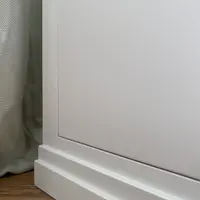 Close view of the plain painted end panel of the pedestal and its two-step moulded plinth, set against a sage linen curtain with oak flooring below