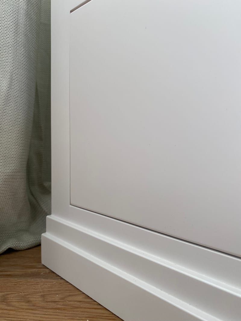 Close view of the plain painted end panel of the pedestal and its two-step moulded plinth, set against a sage linen curtain with oak flooring below
