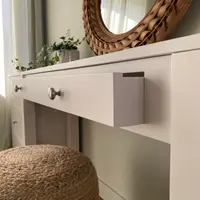 Angled view along the front of the dressing table with the centre drawer eased open to show the painted drawer side, plus brushed nickel knobs, the rattan mirror and jute pouffe