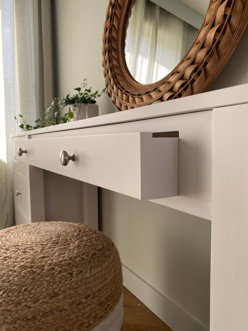 Angled view along the front of the dressing table with the centre drawer eased open to show the painted drawer side, plus brushed nickel knobs, the rattan mirror and jute pouffe