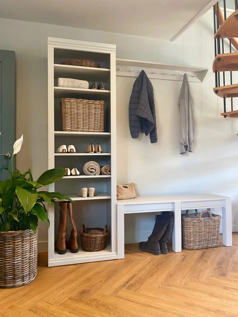 White painted modular boot room with a tall open shoe shelf unit holding boots and baskets, a wall rail of coat pegs with two coats hanging, and a low bench with baskets beneath