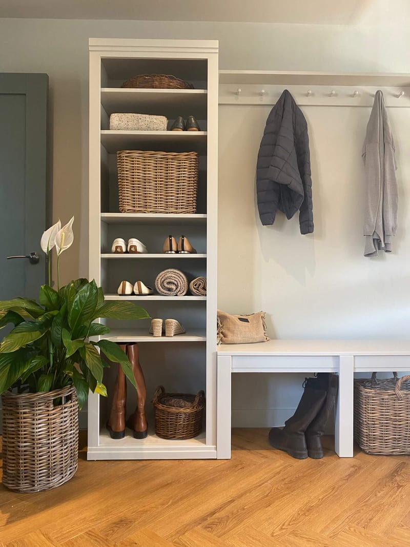 Straight-on view of the white boot room combination, its tall shelf unit stacked with baskets, shoes and rolled throws beside a peg rail holding two coats and a bench with black boots below