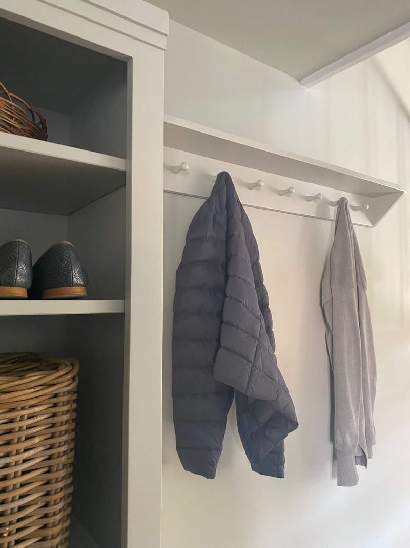 Close view of the wall-mounted peg rail with its row of turned white pegs, a charcoal quilted jacket and a grey knit hanging beside the shelf unit of shoes and a rattan basket